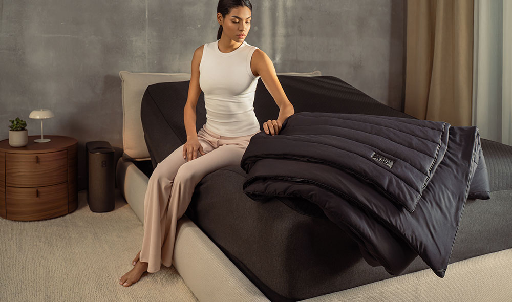 A woman relaxes in a brightly lit bedroom, sitting on a freshly made Eight Sleep Pod 5 bed.